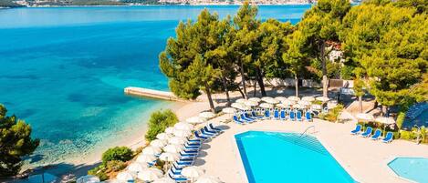 A resort pool area with clear blue water, lounge chairs, and umbrellas, surrounded by trees and a view of the sea.