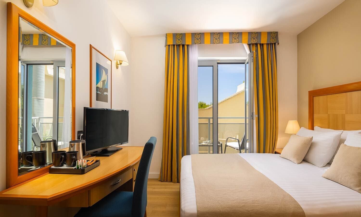 A hotel room with a bed, a desk with a computer, a chair, a television, and a window with curtains.