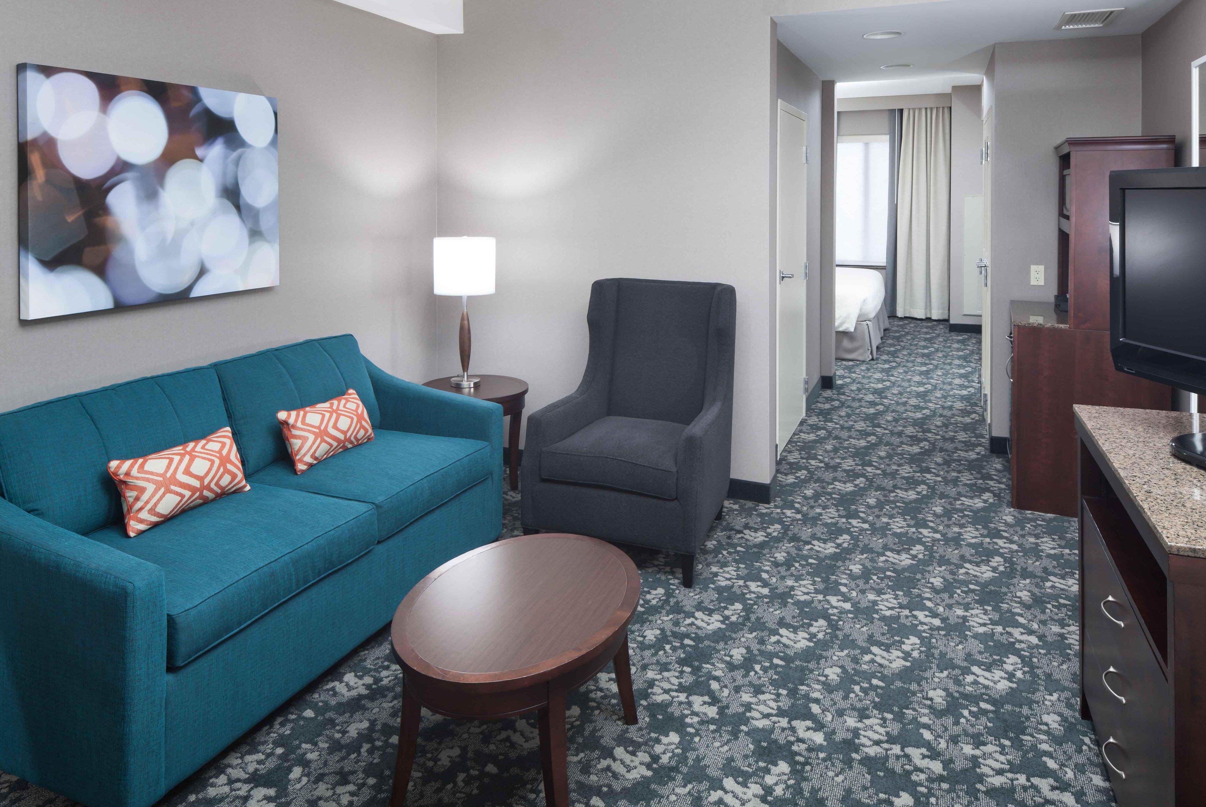 A hotel room with a teal sofa, a small round table, a grey armchair, a TV, and a painting on the wall.