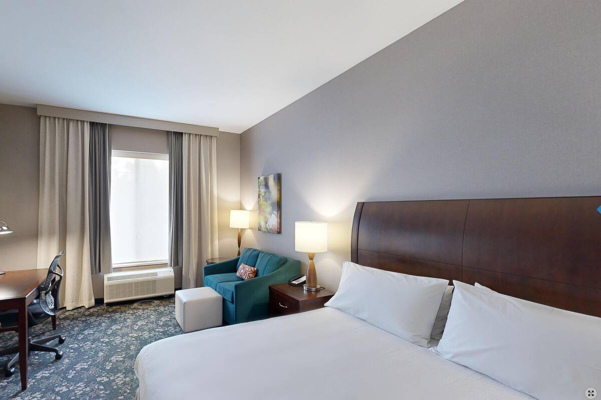 A hotel room with a bed, a desk, a chair, a window with curtains, and a painting on the wall.