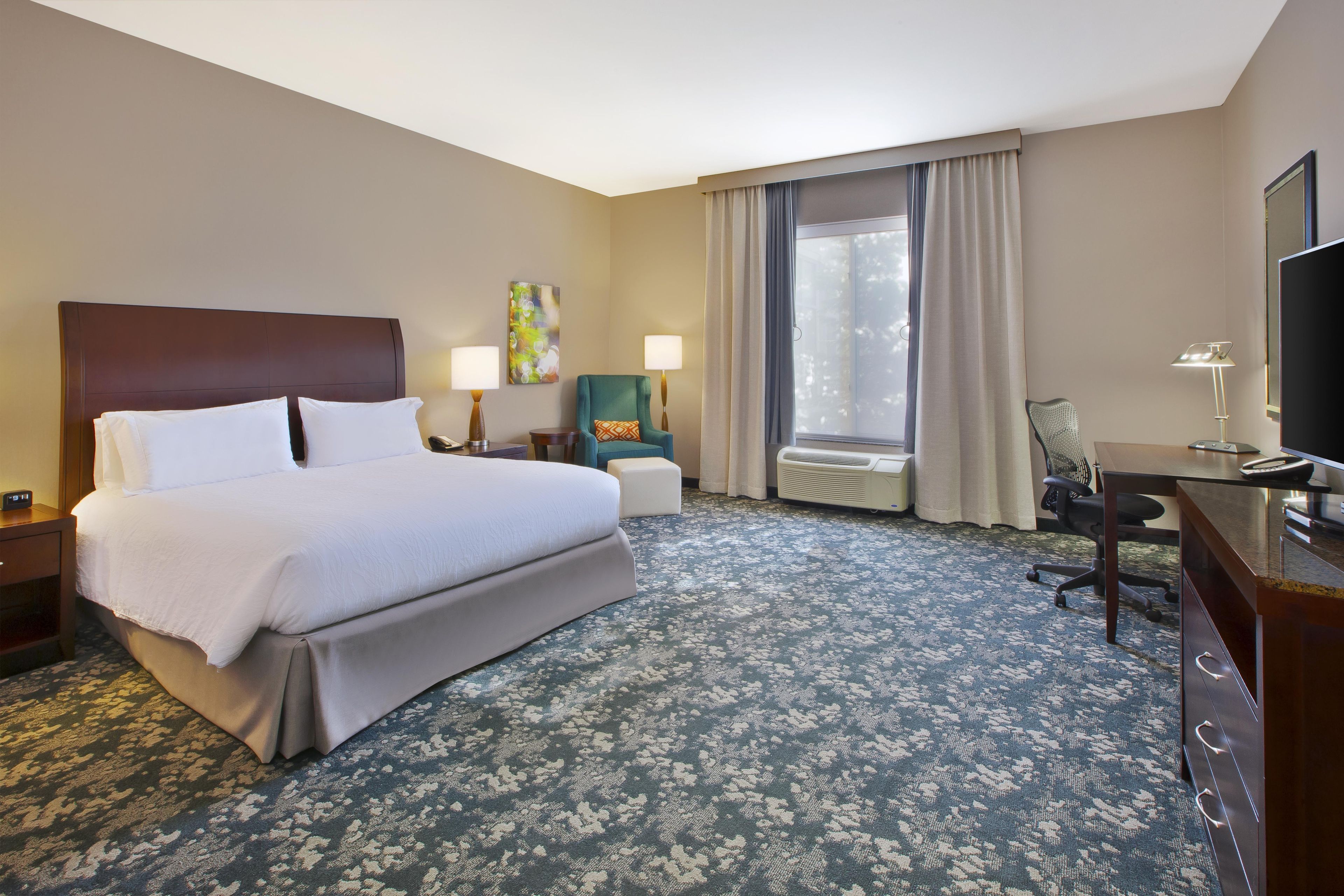 A hotel room with a large bed, a desk, a chair, a TV, and a window with curtains.