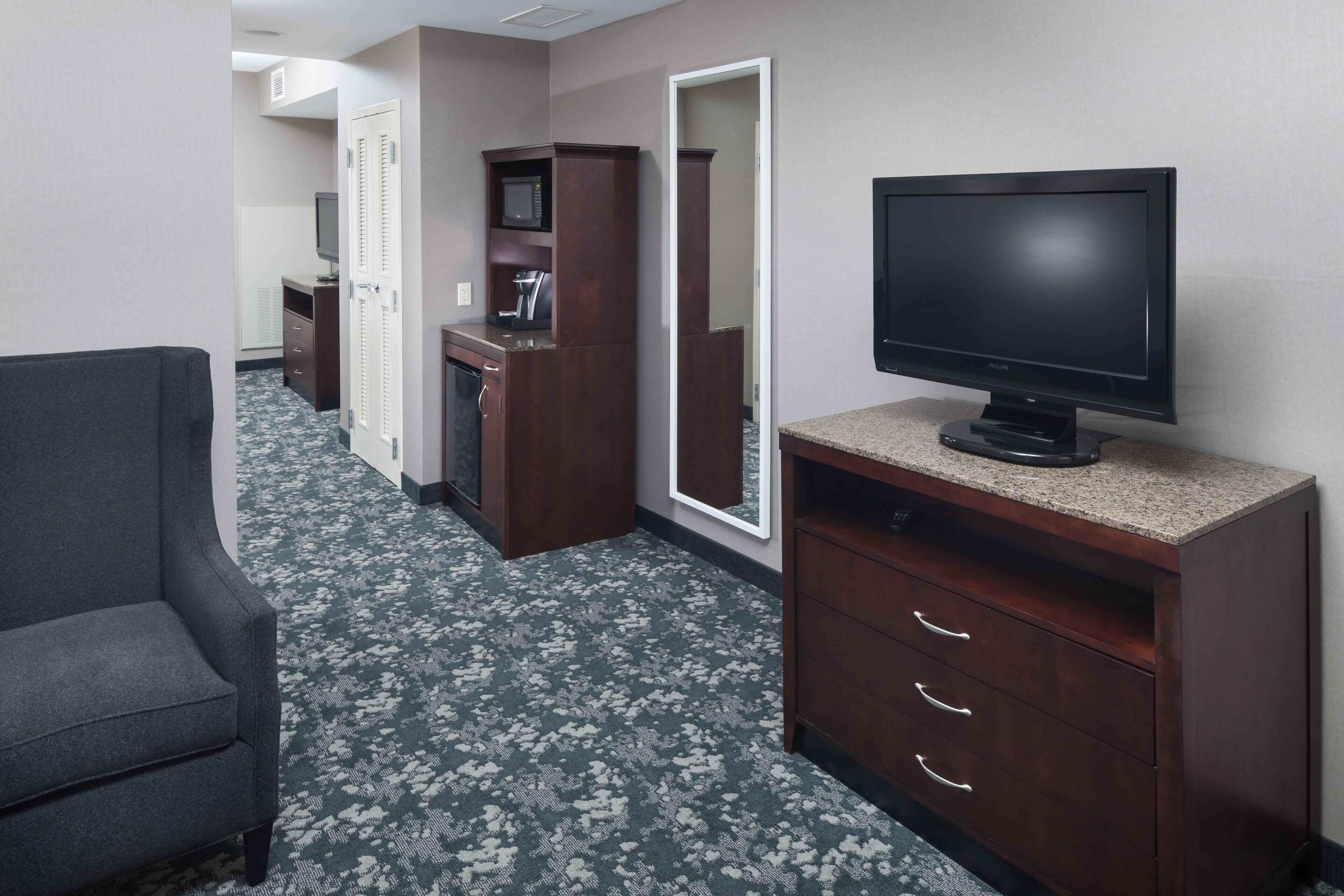 A hotel room with a dark wood dresser, a television, a gray armchair, and a patterned carpet.