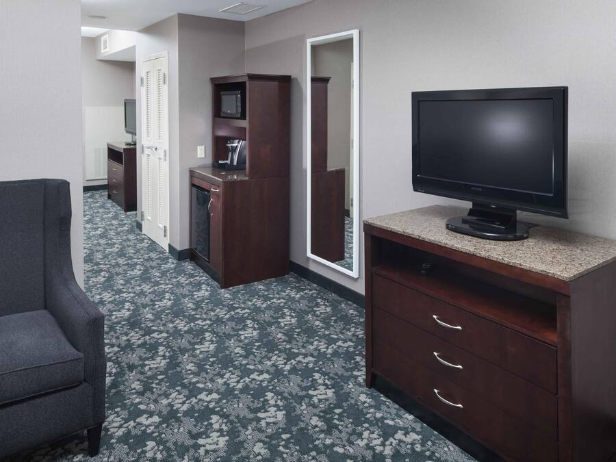 A hotel room with a dark wood dresser, a television, a gray armchair, and a patterned carpet.