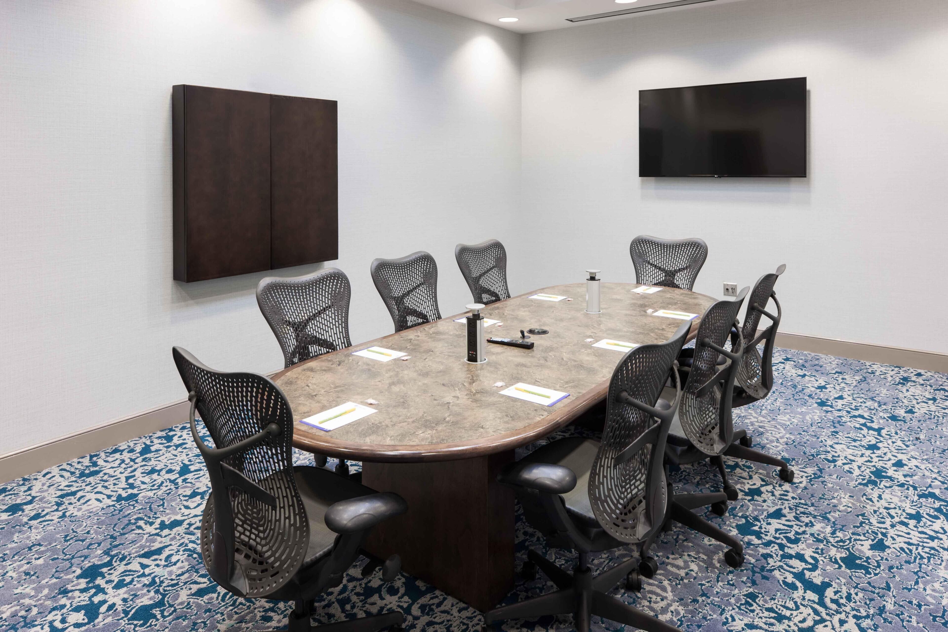 A conference room with a large table, eight mesh chairs, a flat-screen TV, and two dark panels on the walls.