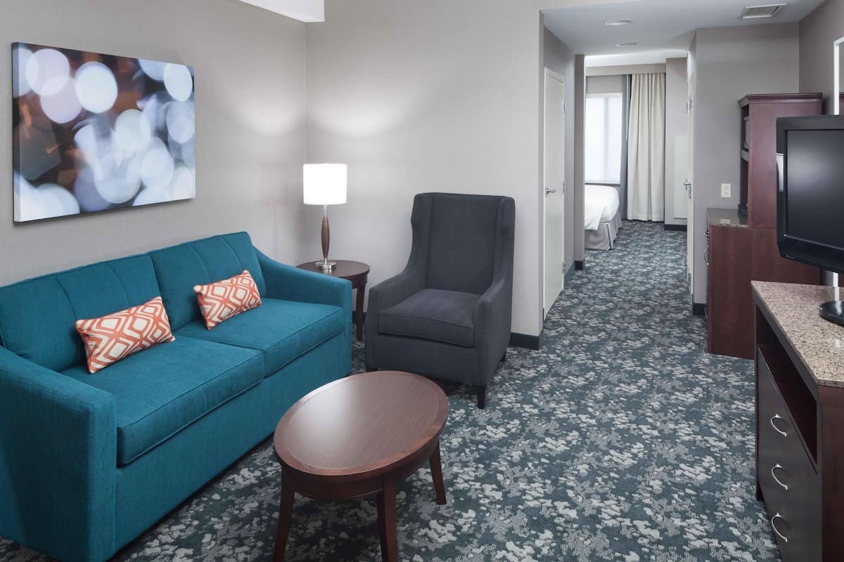 A hotel room with a teal sofa, a small round table, a grey armchair, a TV, and a painting on the wall.