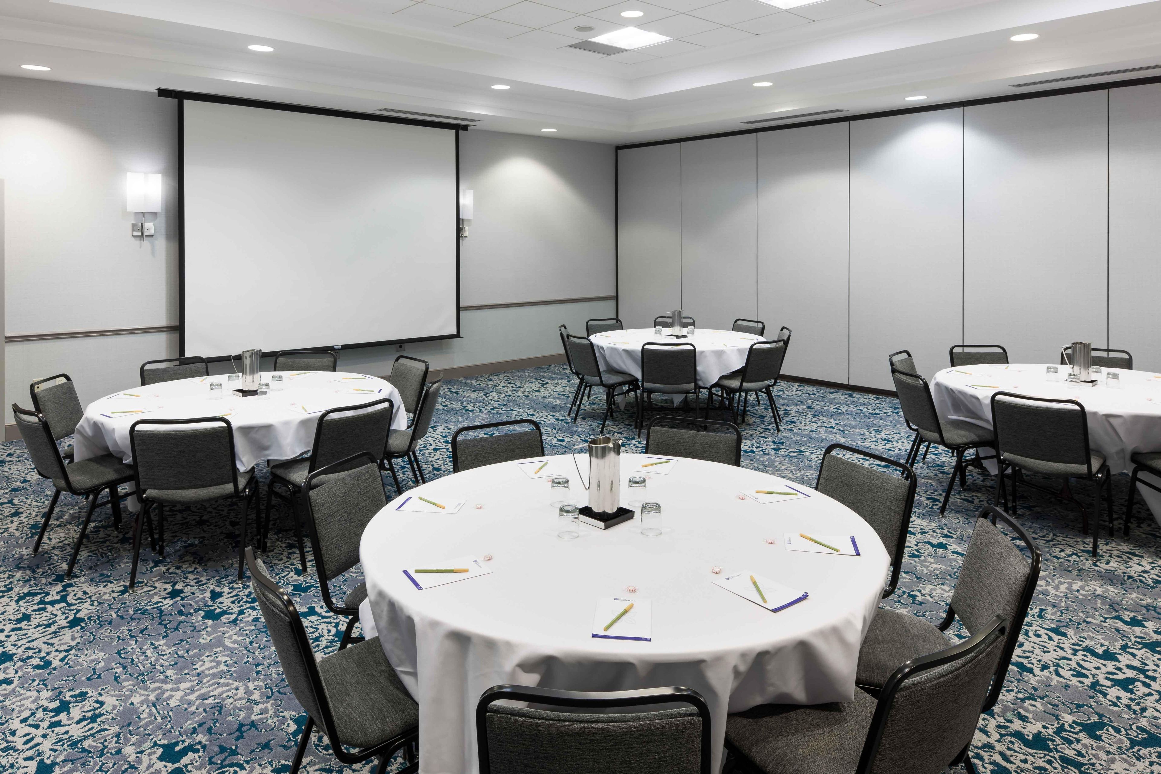 A conference room with multiple round tables, each with chairs, white tablecloths, and a projector screen.