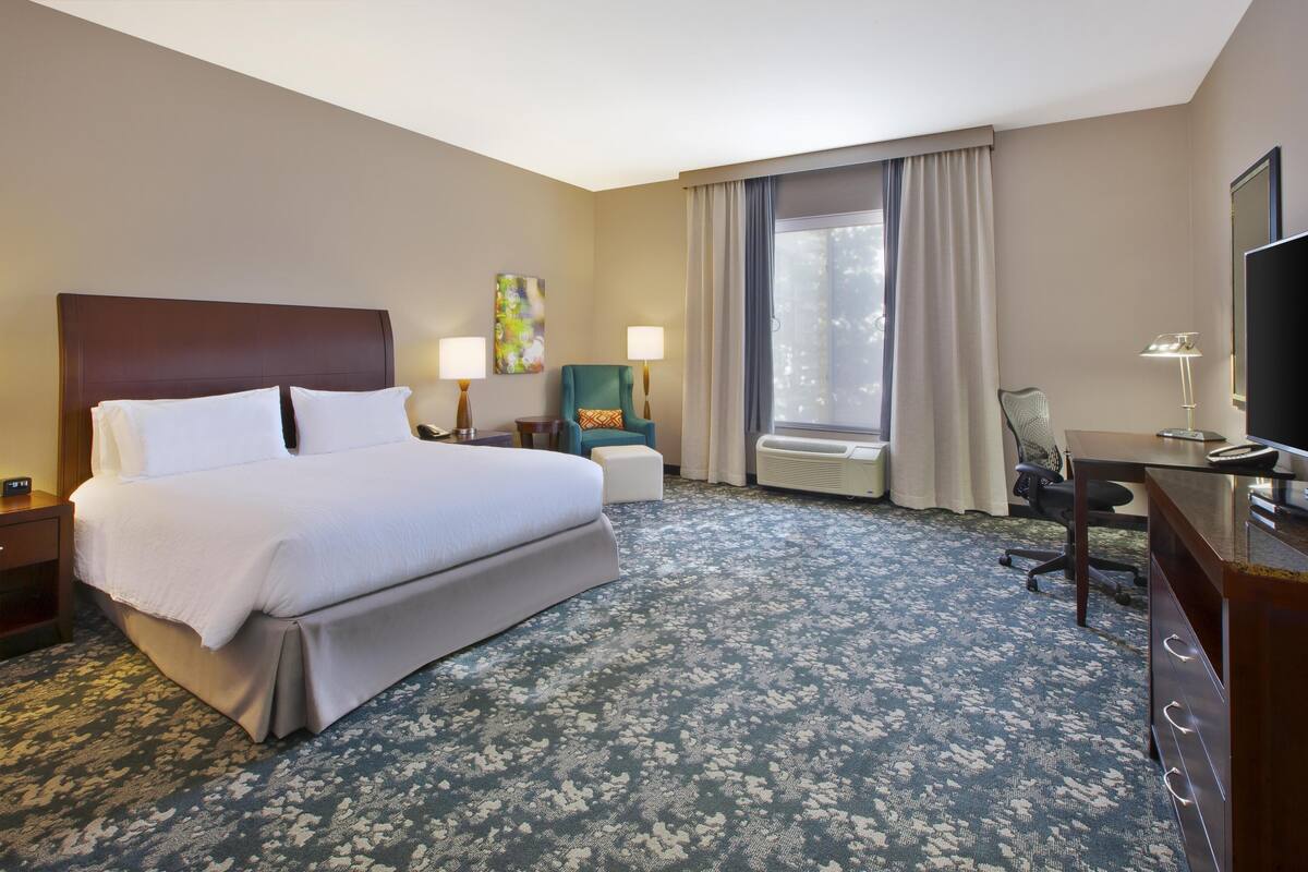 A hotel room with a large bed, a desk, a chair, a TV, and a window with curtains.