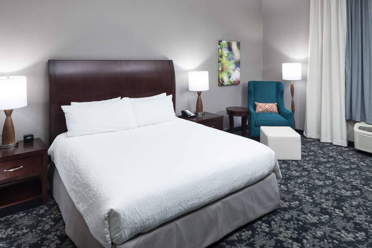 A hotel room with a large bed, bedside tables, a chair, a small table, and a wall-mounted lamp.