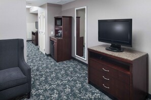 Un couloir d’hôtel avec un fauteuil gris, un bureau en bois sur lequel se trouve une télévision et un four à micro-ondes.