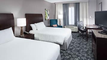 A hotel room with two beds, a desk with a television, a chair, a lamp, and a window with curtains.