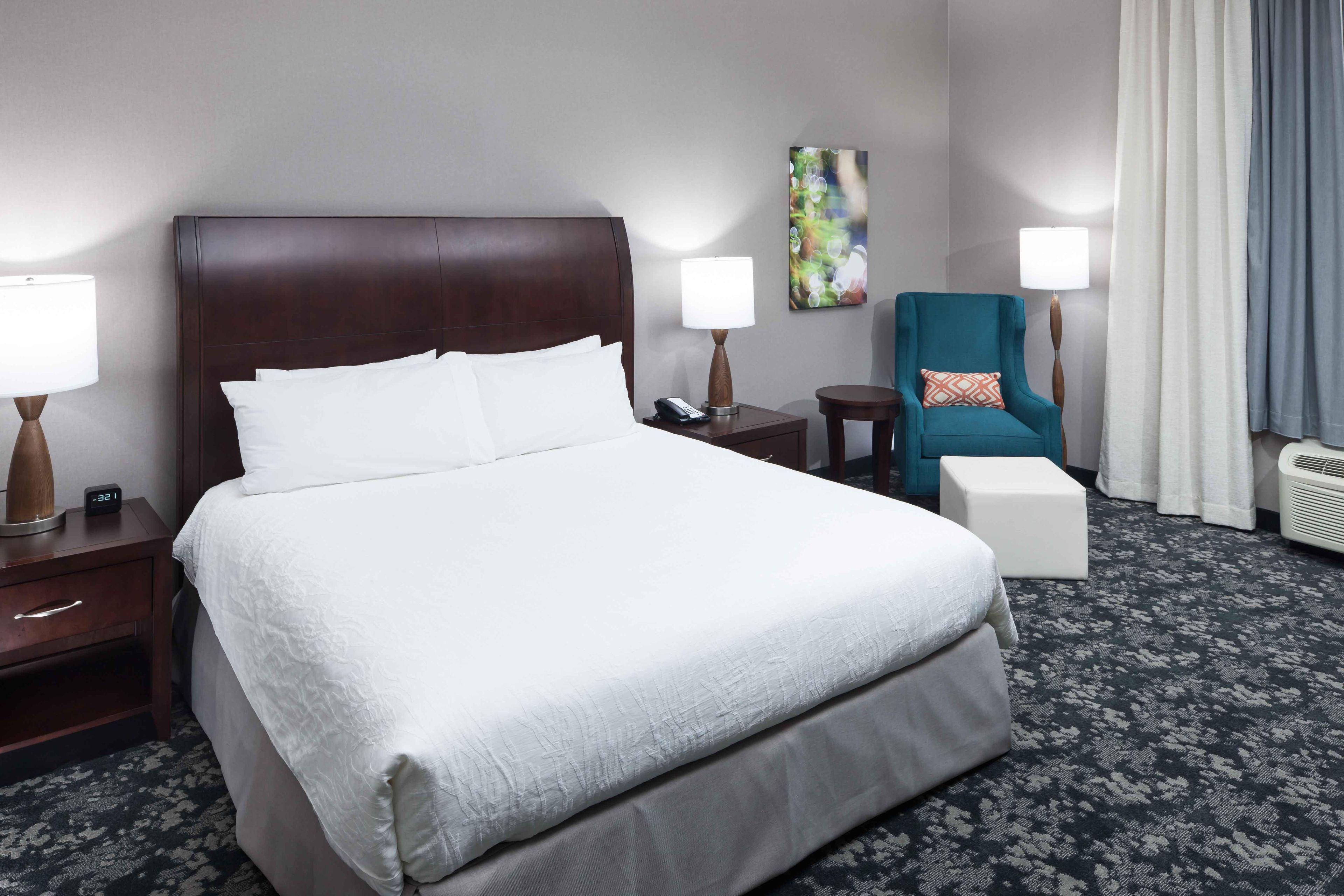 Una habitación de hotel con una cama grande, mesitas de noche, una silla, una pequeña mesa y una lámpara fijada a la pared.