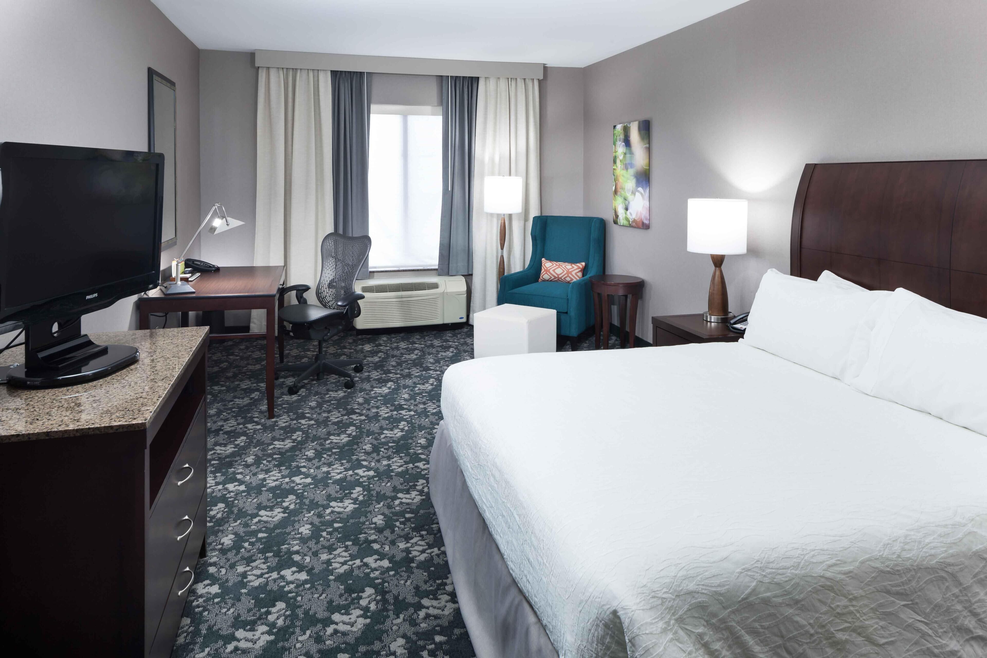 A hotel room with a bed, desk, TV, chair, and a window with curtains.