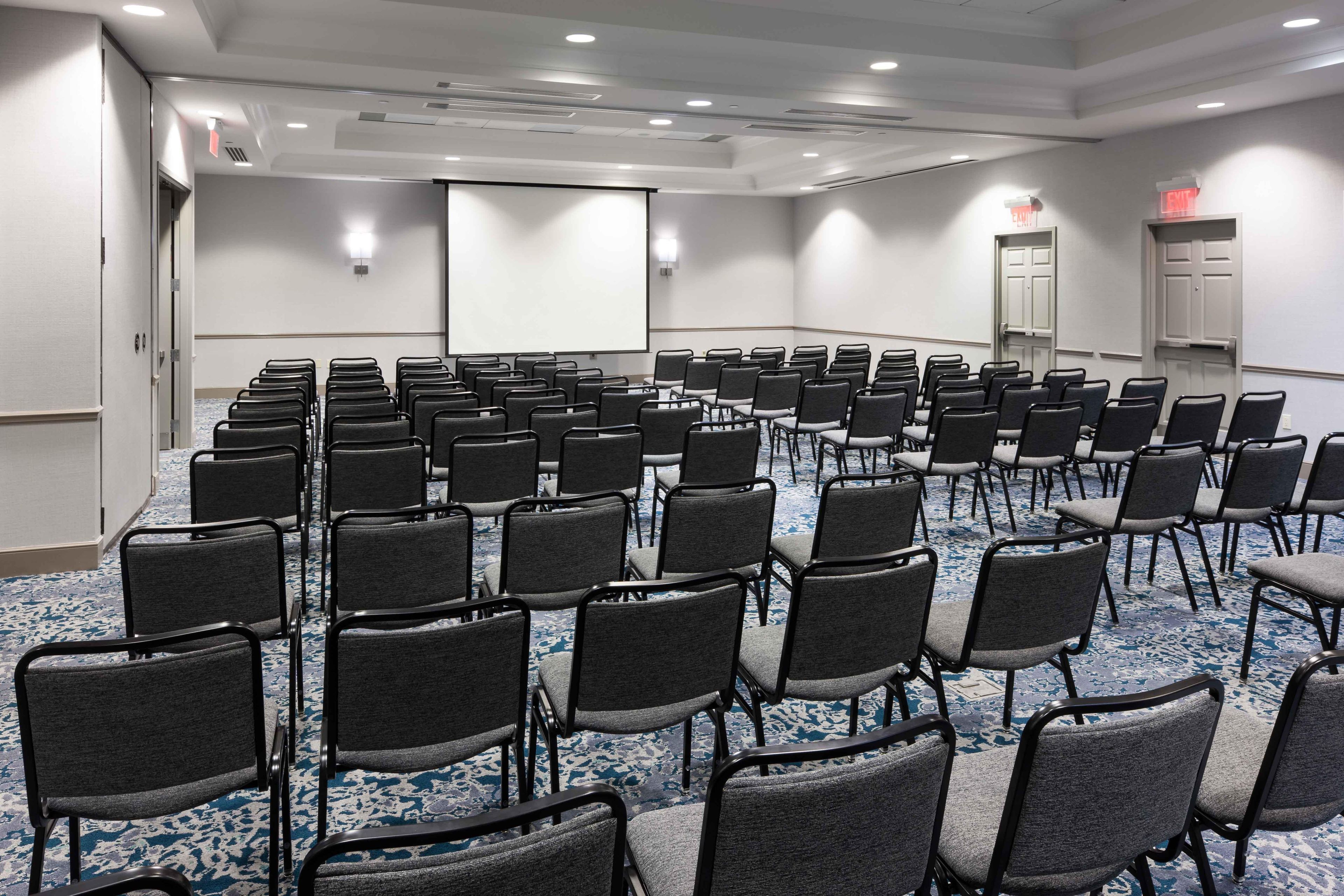 Un salón de conferencias con filas de sillas, una pantalla de proyector y una puerta de salida.