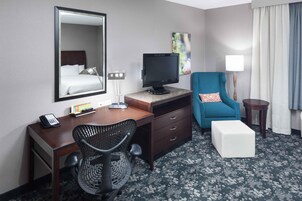 Habitación de hotel con cama, escritorio, silla, televisor y una mesita de noche con lámpara.