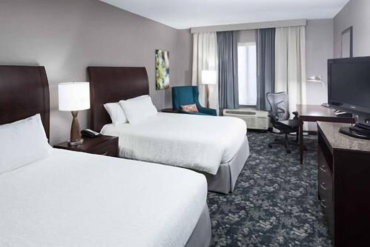 A hotel room with two beds, a desk, a chair, a TV, and a window with curtains.
