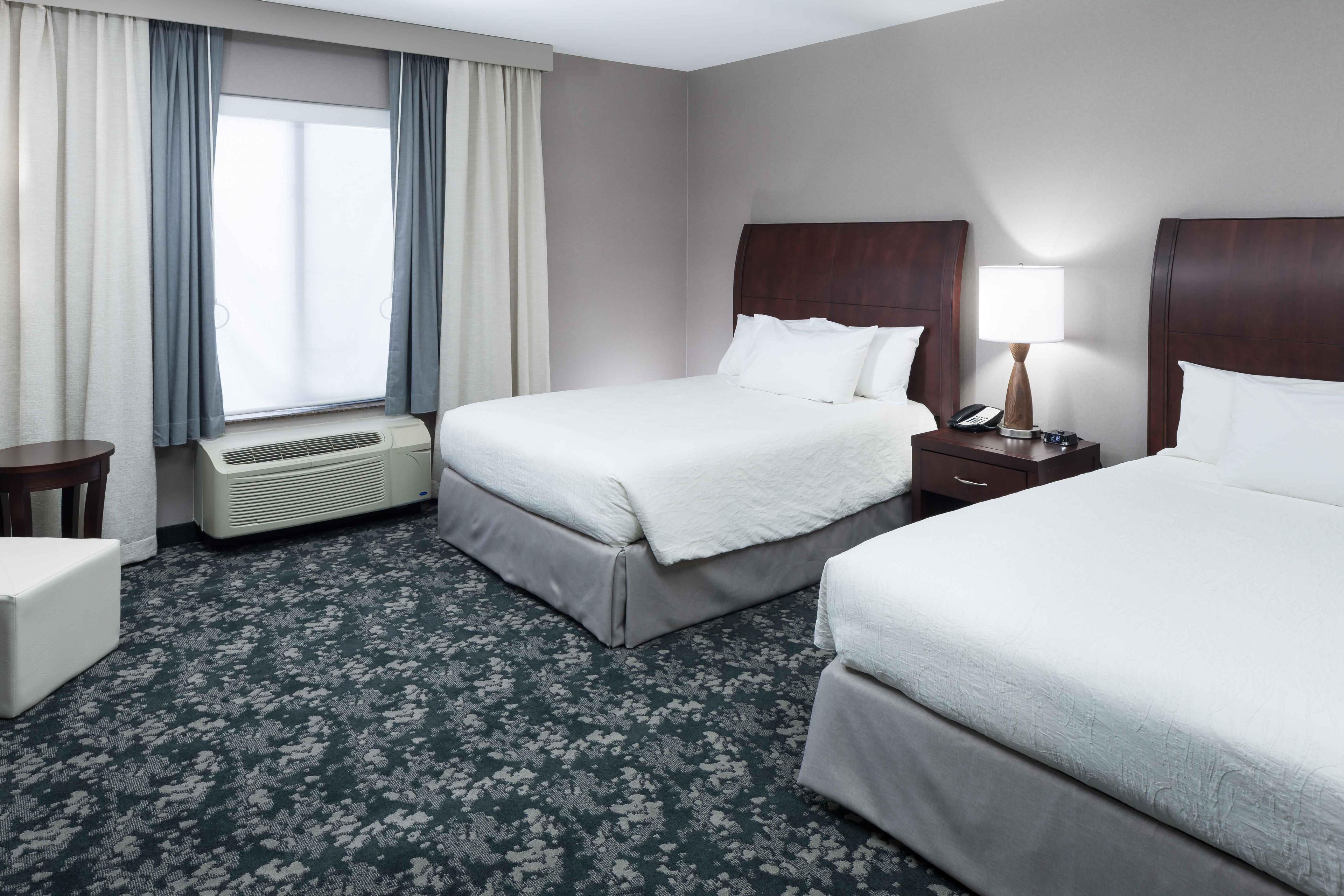 Quarto de hotel com duas camas, uma mesinha de cabeceira com abajur, telefone e relógio de parede.