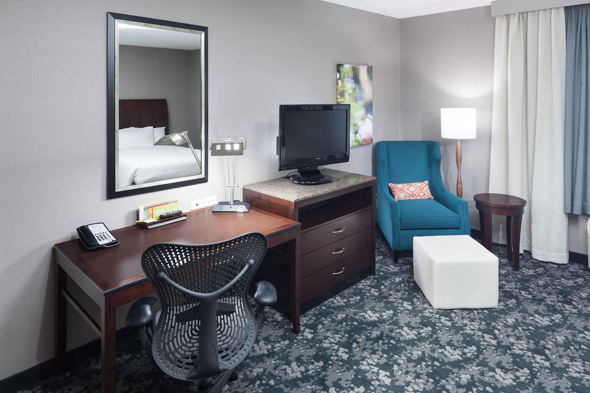 A hotel room with a bed, a desk with a computer, a chair, a TV, a lamp, and a small table.