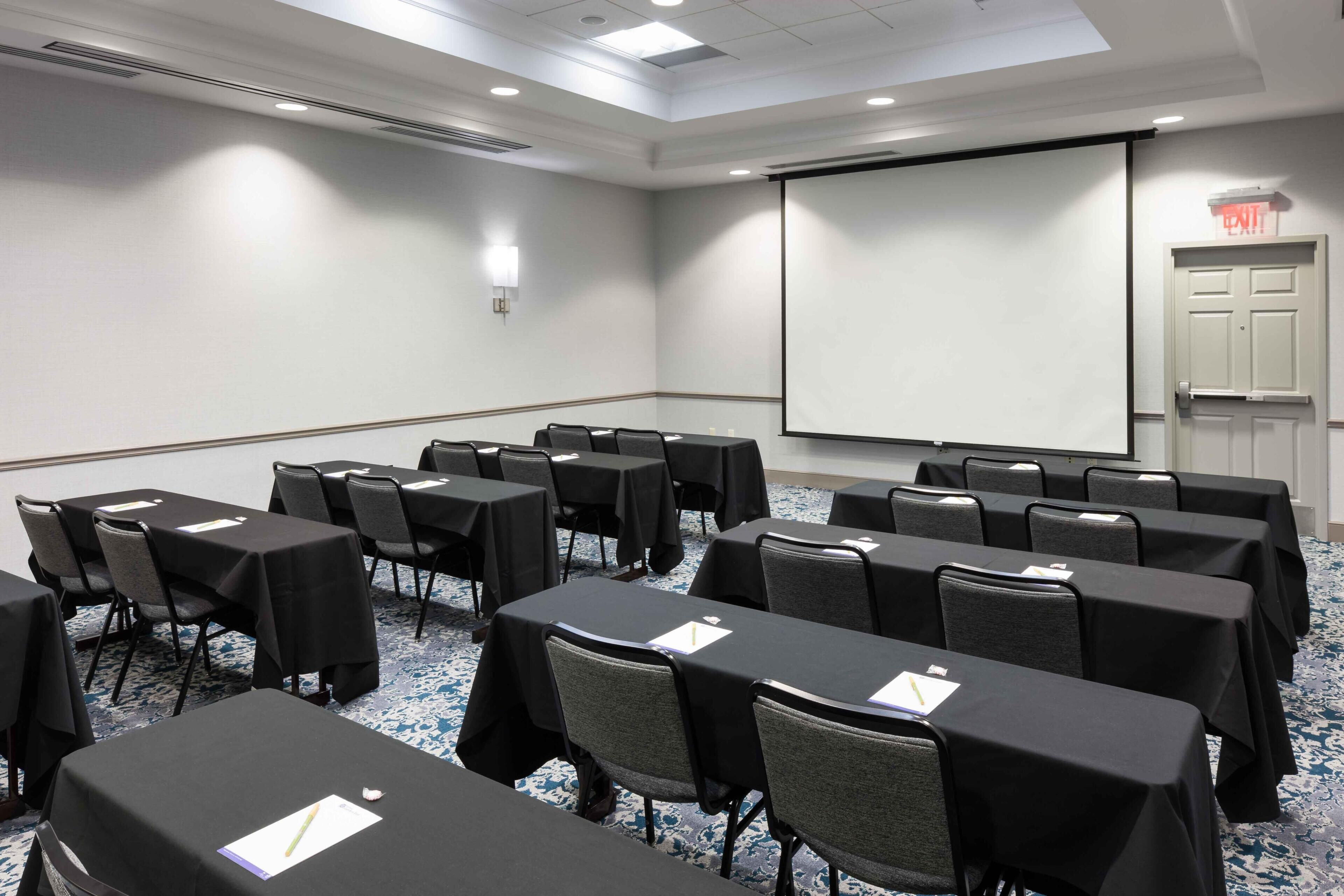 A conference room with tables, chairs, a projector screen, and an exit door.