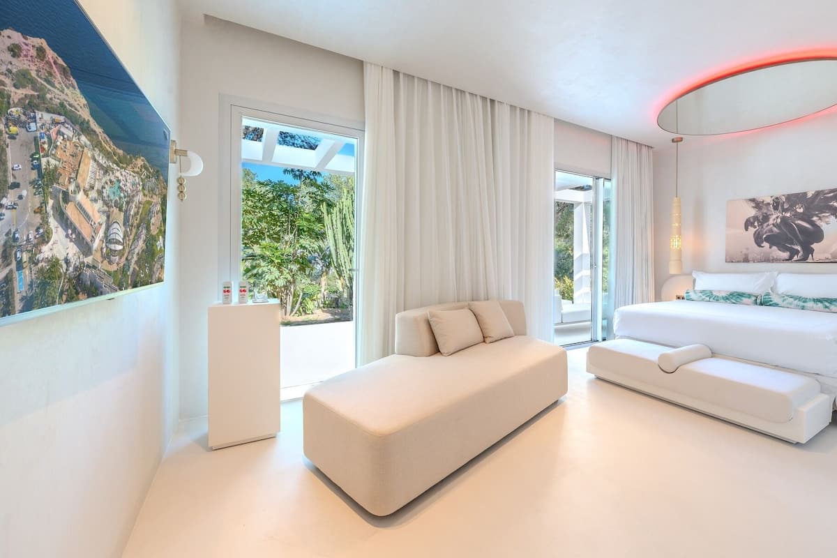 A modern hotel room with a large bed, a sofa, a dining table, and a view of a coastal area.