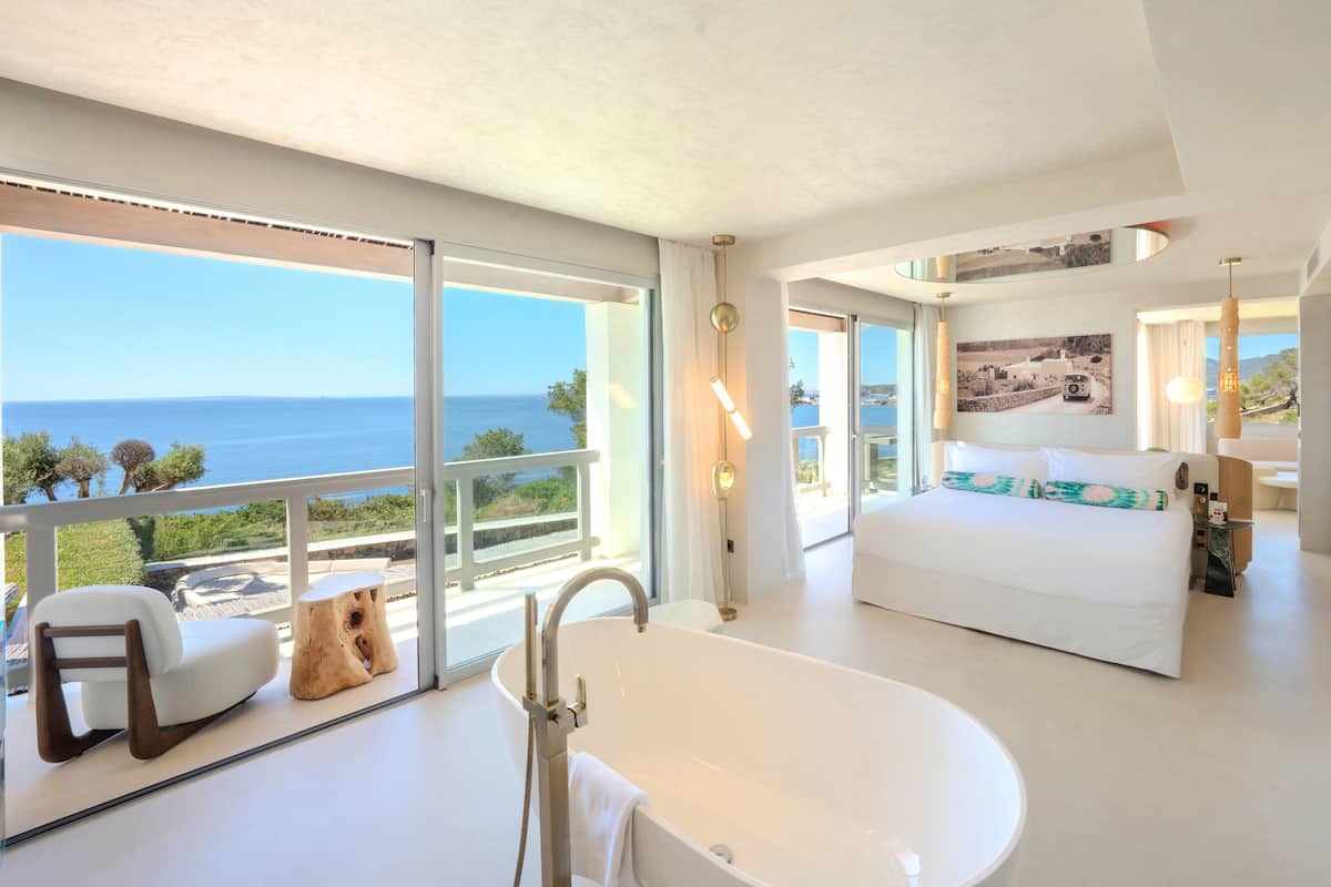 A modern hotel room with a large window, a bathtub, a bed, and a balcony with a view of the sea.