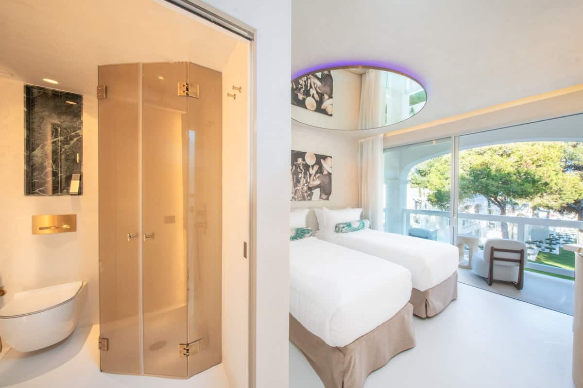 A modern hotel room with a glass shower enclosure, two beds, a large window with a view, and a minibar.