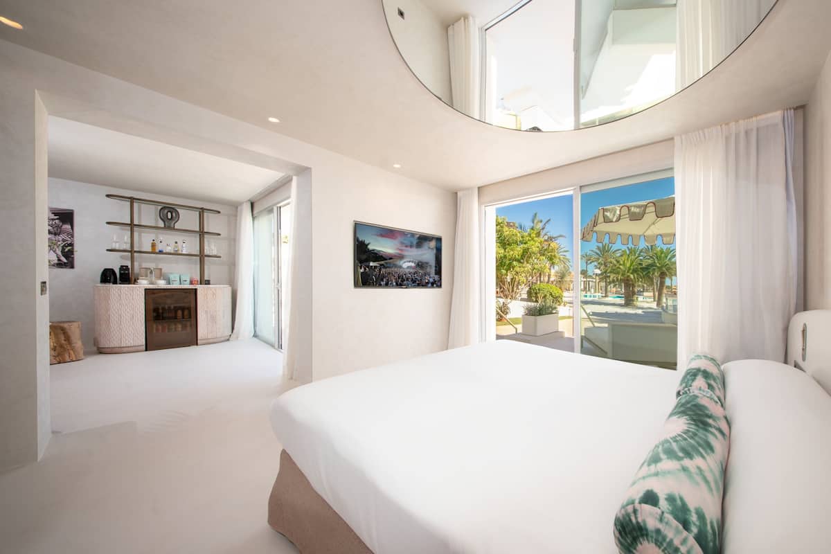 A modern hotel room with a large bed, a seating area, a kitchenette, and a view of the outdoors.