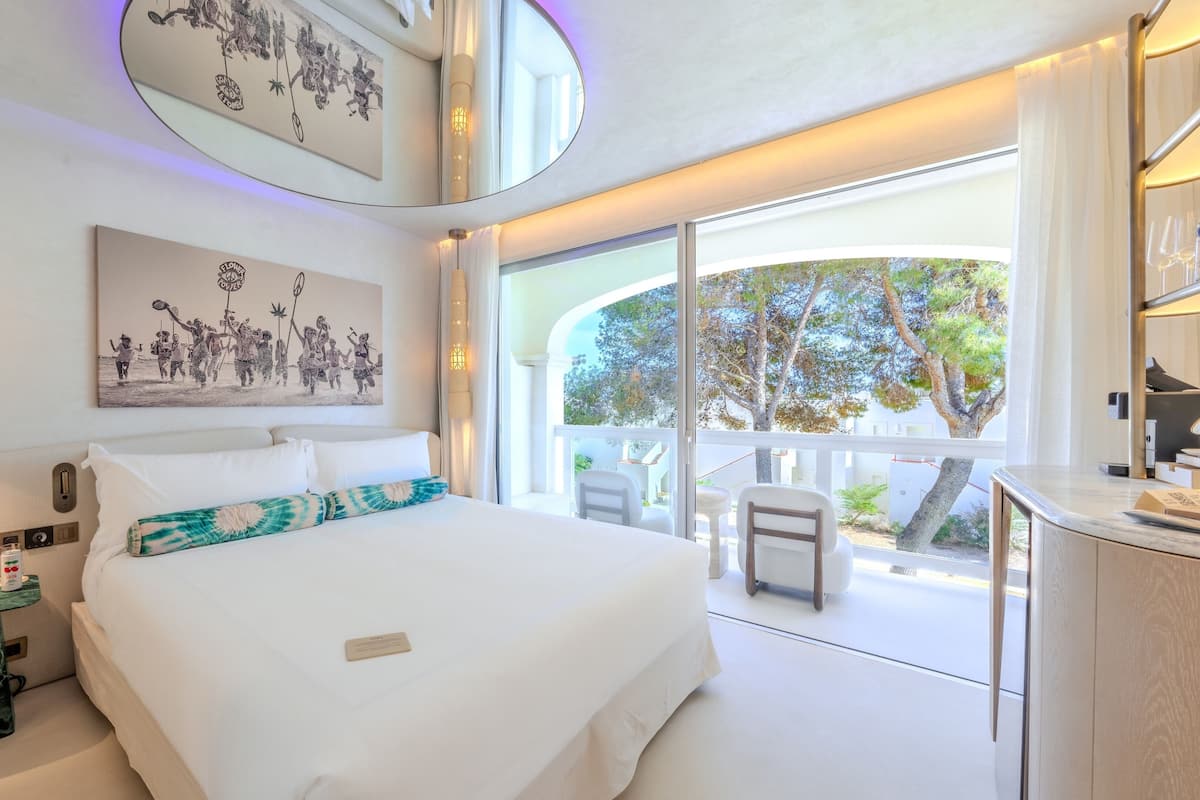 A modern hotel room with a large bed, a glass table, a balcony with a view, and a ceiling light fixture.