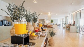 Un moderno vestíbulo de hotel con una zona de bufé que ofrece frutas frescas, una estación de café y plantas decorativas.