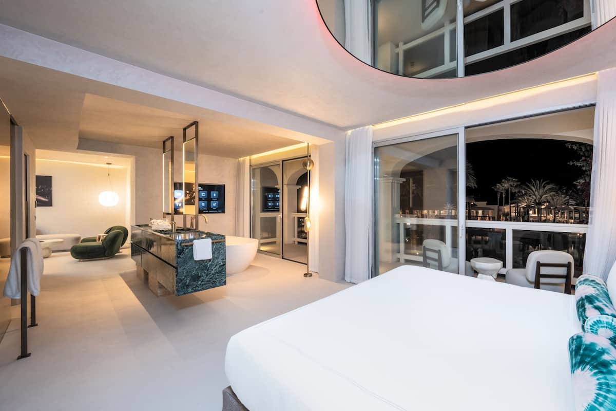 A modern hotel room with a large bed, a bathroom with a bathtub, a seating area with a sofa and chairs, and a balcony with a view of the night sky.