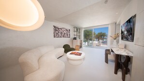 Una sala de estar moderna con un sofá curvo, una mesita redonda con una revista, una silla verde, una mesita auxiliar de madera y un ventanal con vistas a palmeras y una piscina.