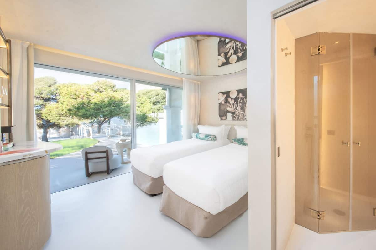 A modern hotel room with two beds, a bathroom with a large mirror and sink, and a view of a courtyard with trees.