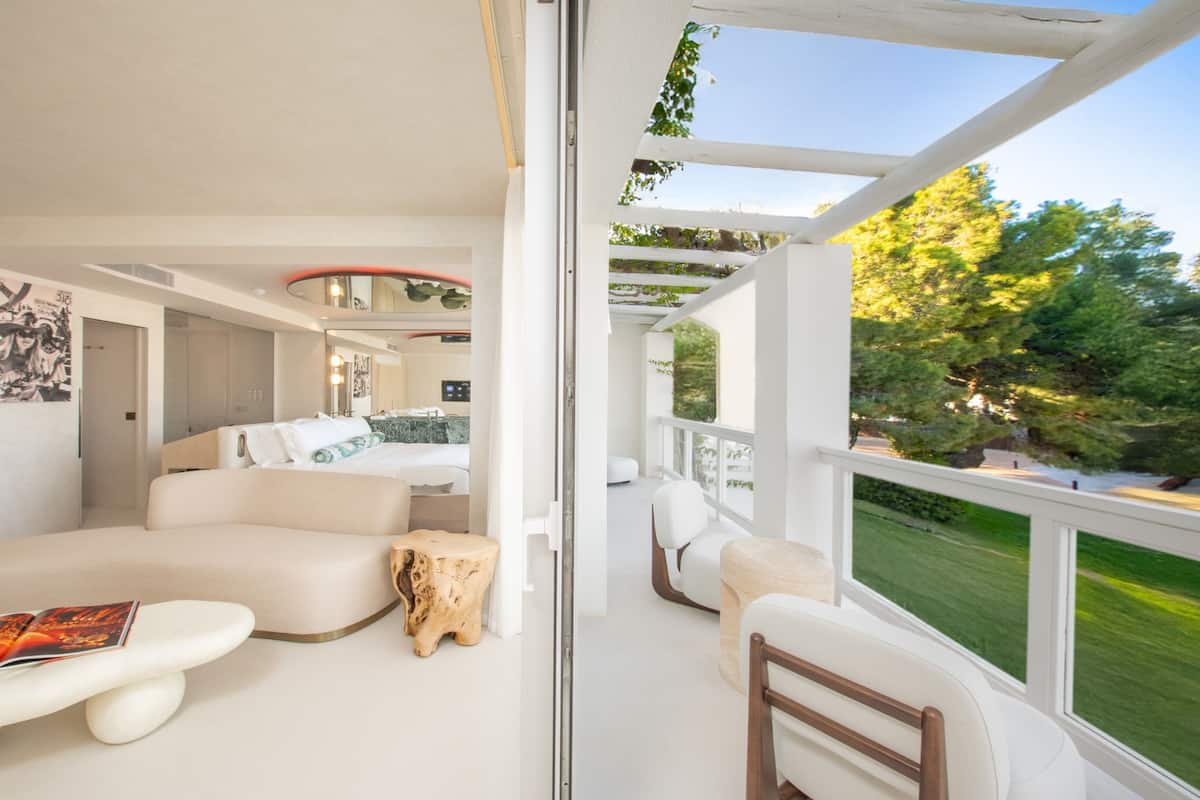 A modern hotel room with a balcony, a bed, a seating area, and a view of the outdoors.