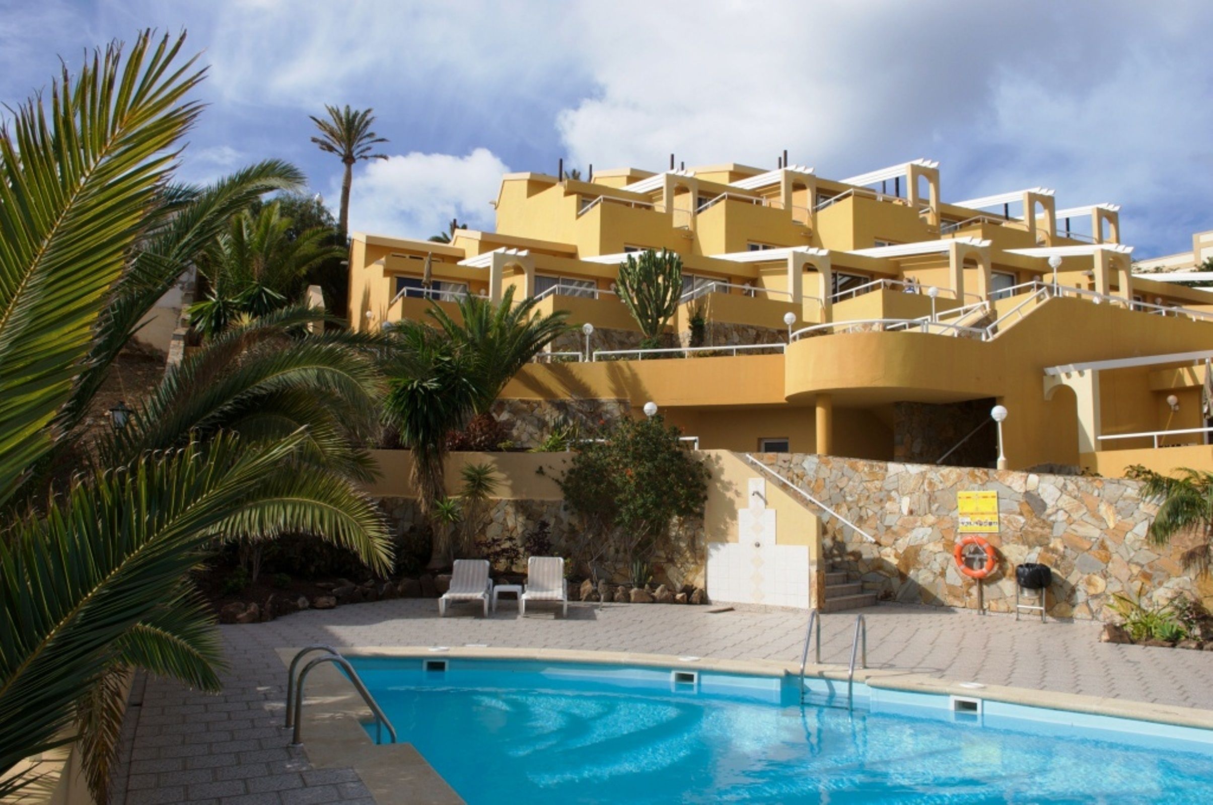 A hotel with a swimming pool, palm trees, and a stone wall.