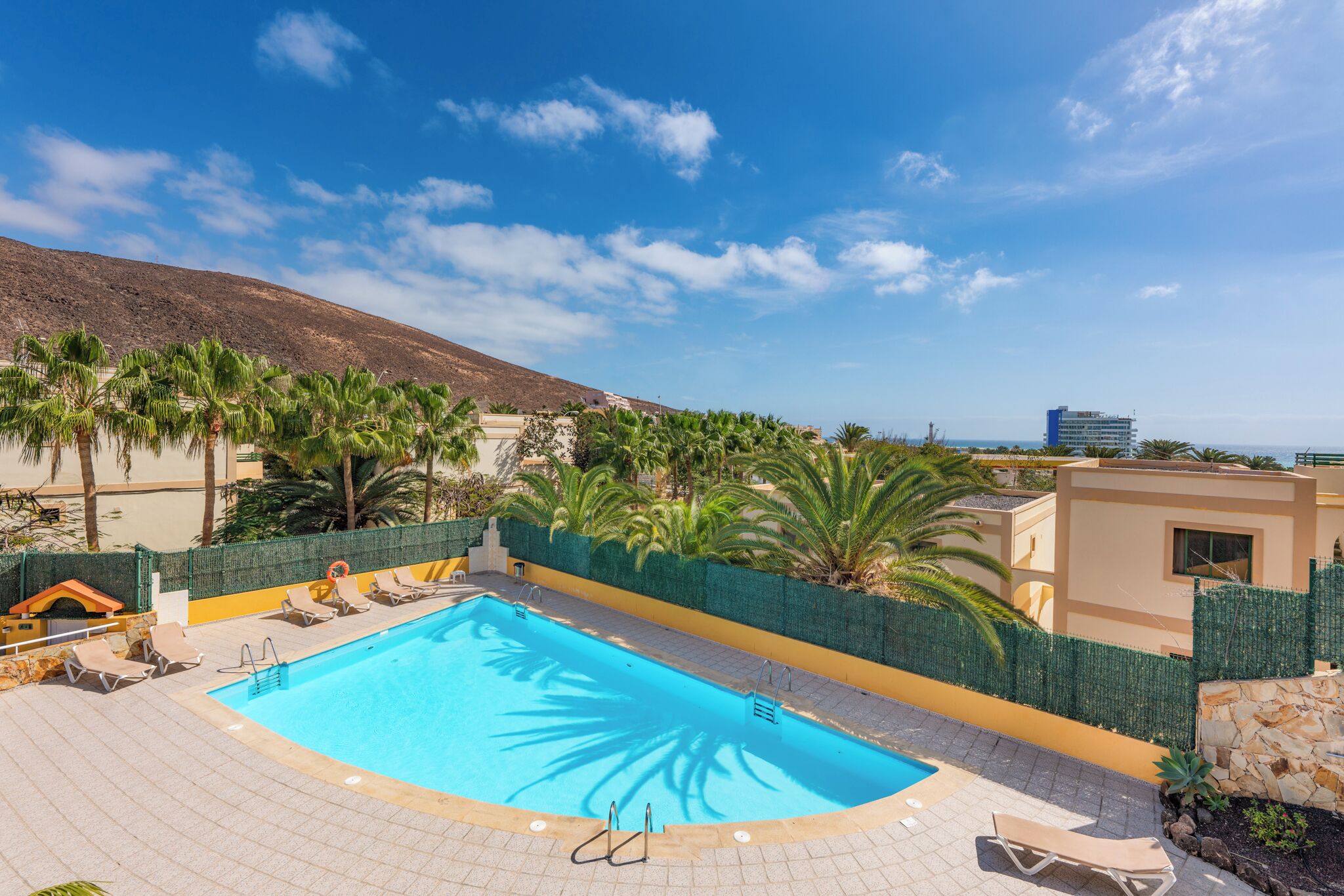 A swimming pool with lounge chairs, a stone wall, and a view of the sea and mountains.