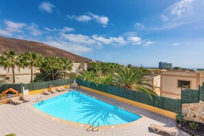 A swimming pool with lounge chairs, a stone wall, and a view of the sea and mountains.