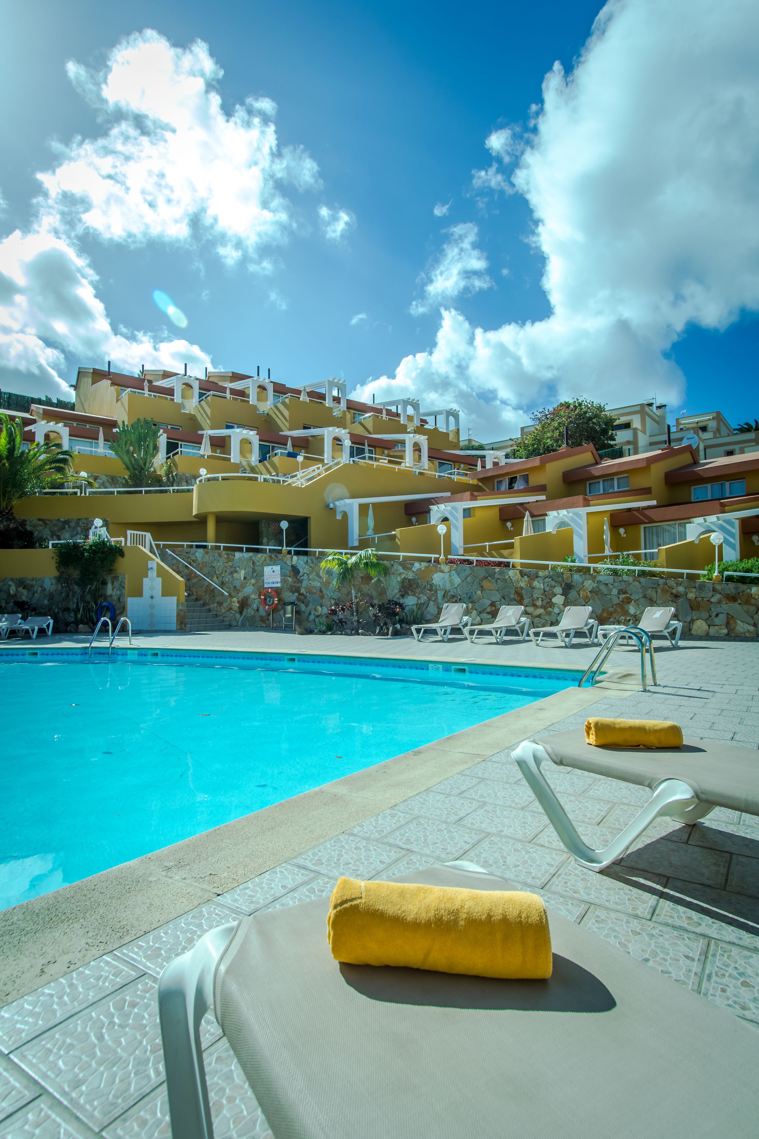A swimming pool with lounge chairs and a yellow towel, surrounded by a building with multiple balconies and a clear blue sky.