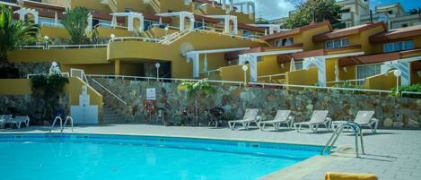 A swimming pool with lounge chairs and a yellow towel, surrounded by a building with multiple balconies and a clear blue sky.