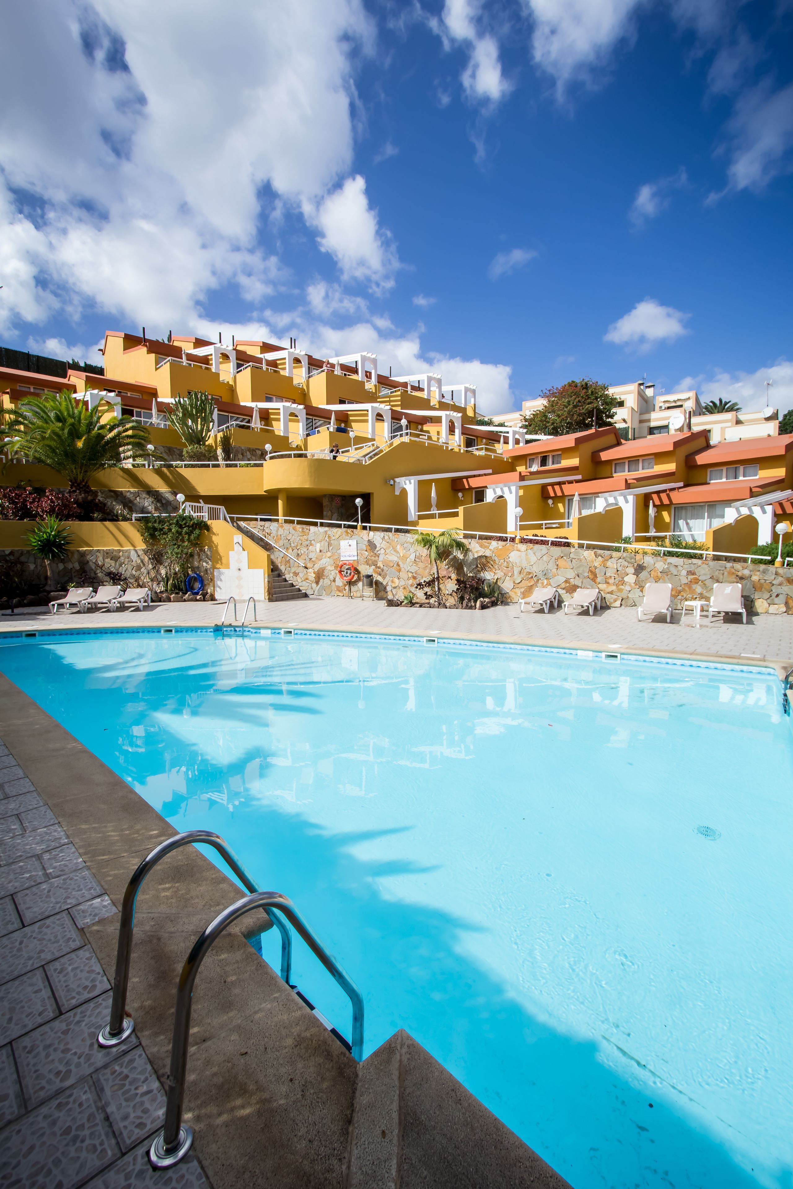 A swimming pool with a clear blue water, surrounded by a stone wall and a metal ladder. In the background, there are yellow buildings with balconies and a clear blue sky.