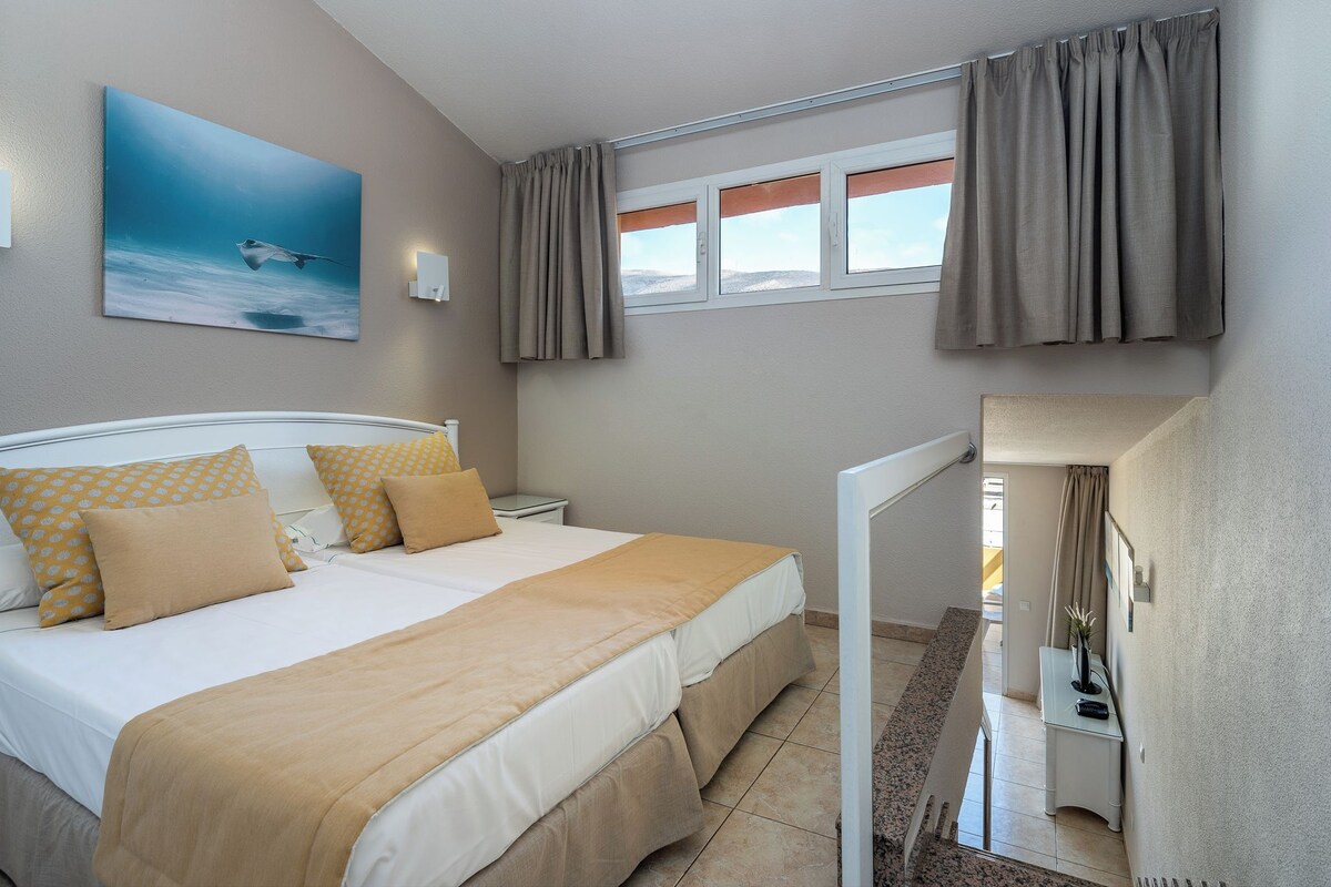 A bedroom with a bed, a nightstand, a bedside table, a window with curtains, and a painting on the wall.
