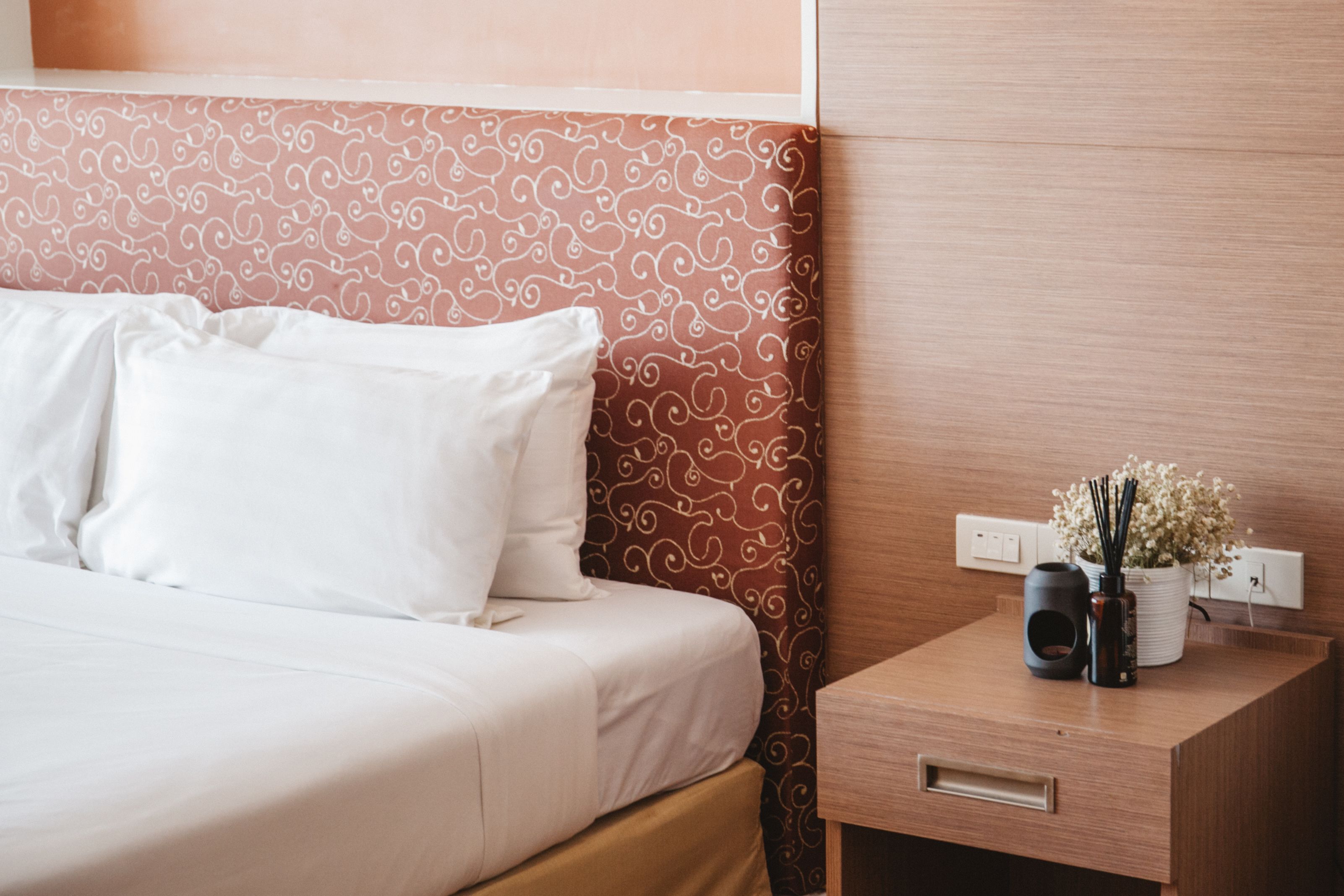 A neatly made bed with white linens, a patterned headboard, and a bedside table with a vase of flowers and a black object.