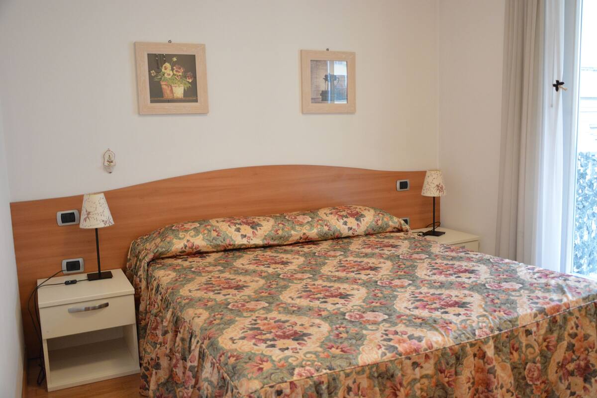 A bedroom with a floral bedspread, wooden headboard, bedside tables with lamps, and two framed pictures on the wall.