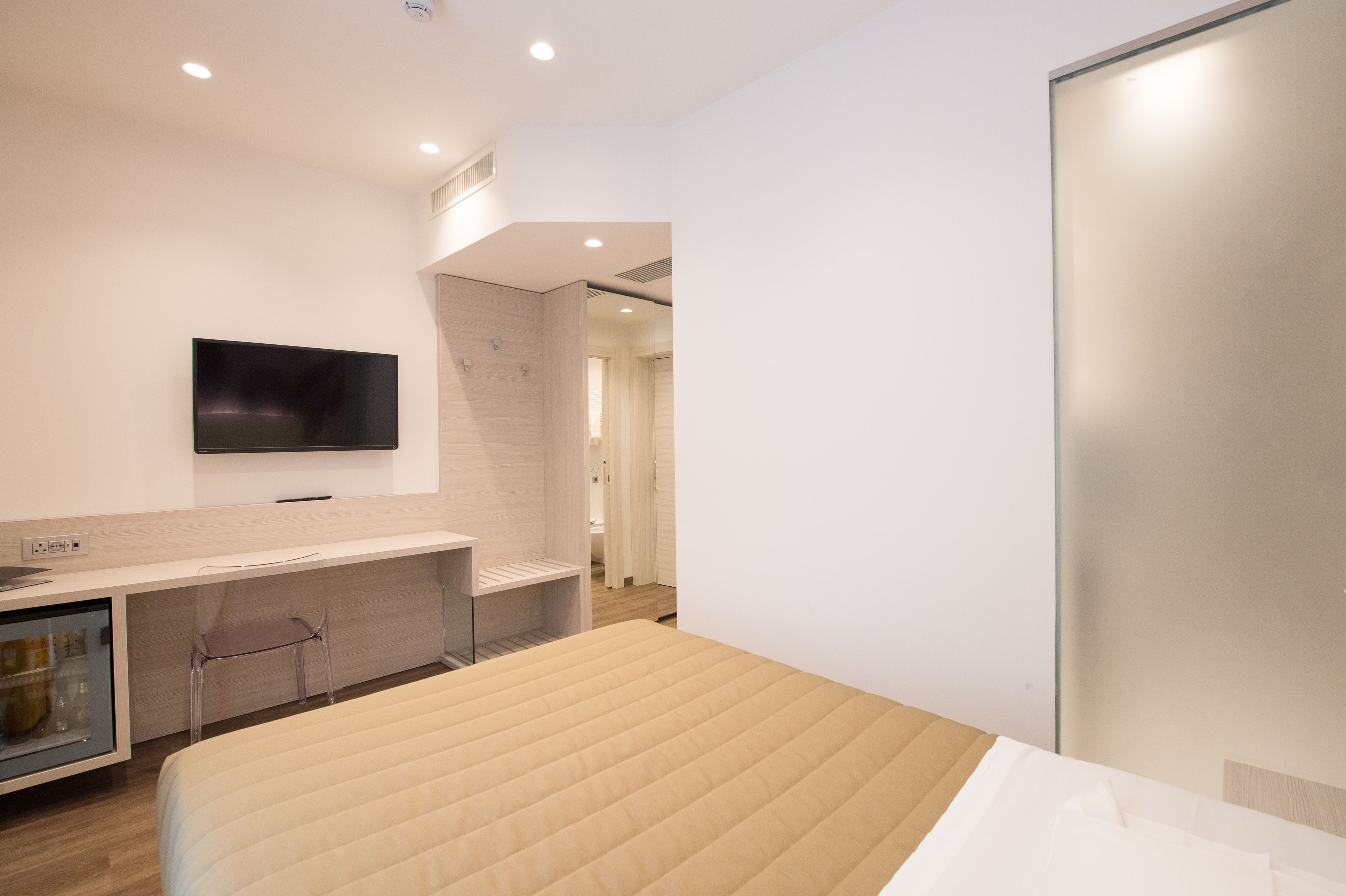 A modern hotel room with a bed, a TV mounted on the wall, a small desk with a chair, and a glass partition.