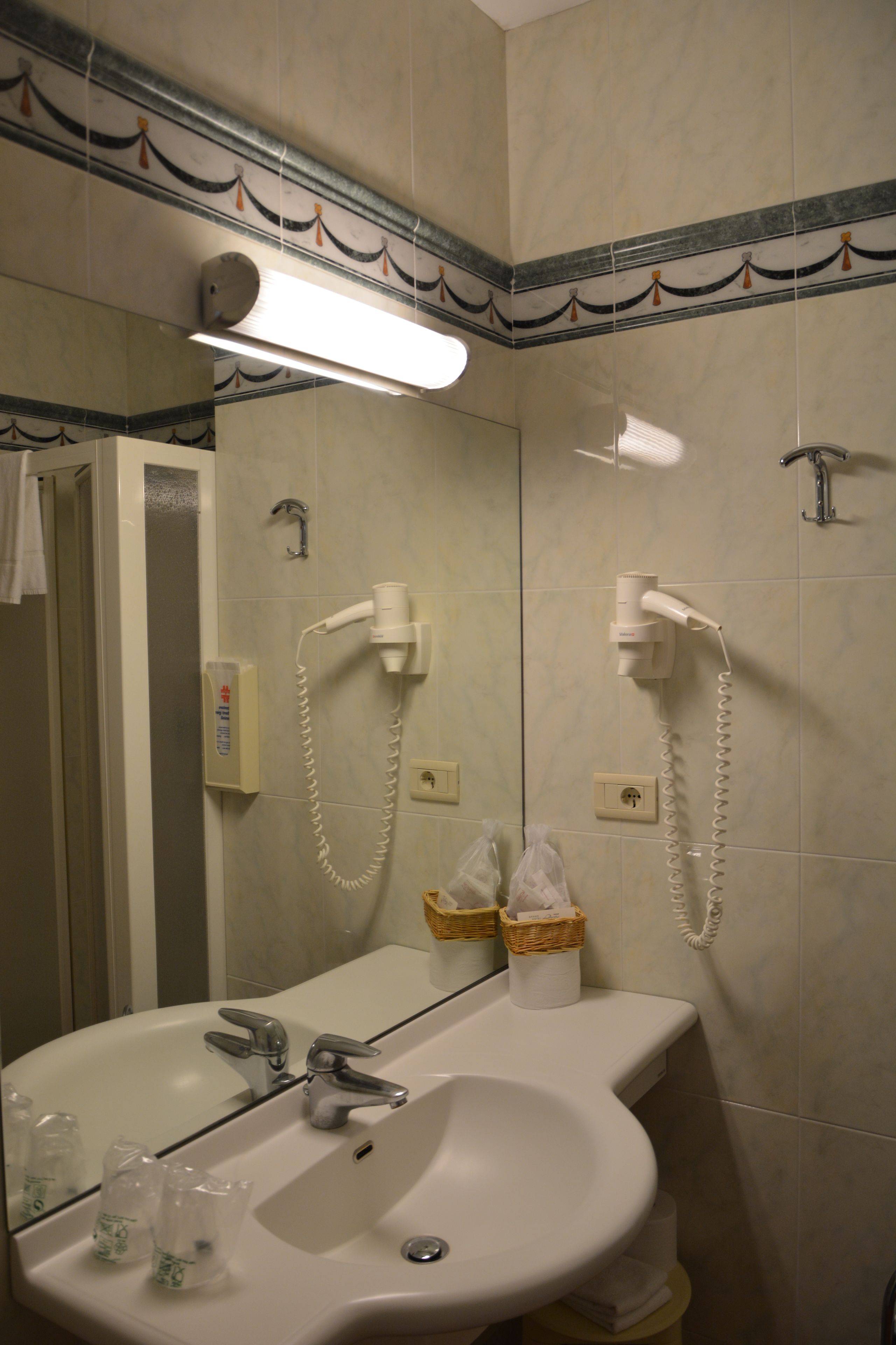 Un baño con lavamanos, espejo y dos secadores de pelo fijados a la pared.