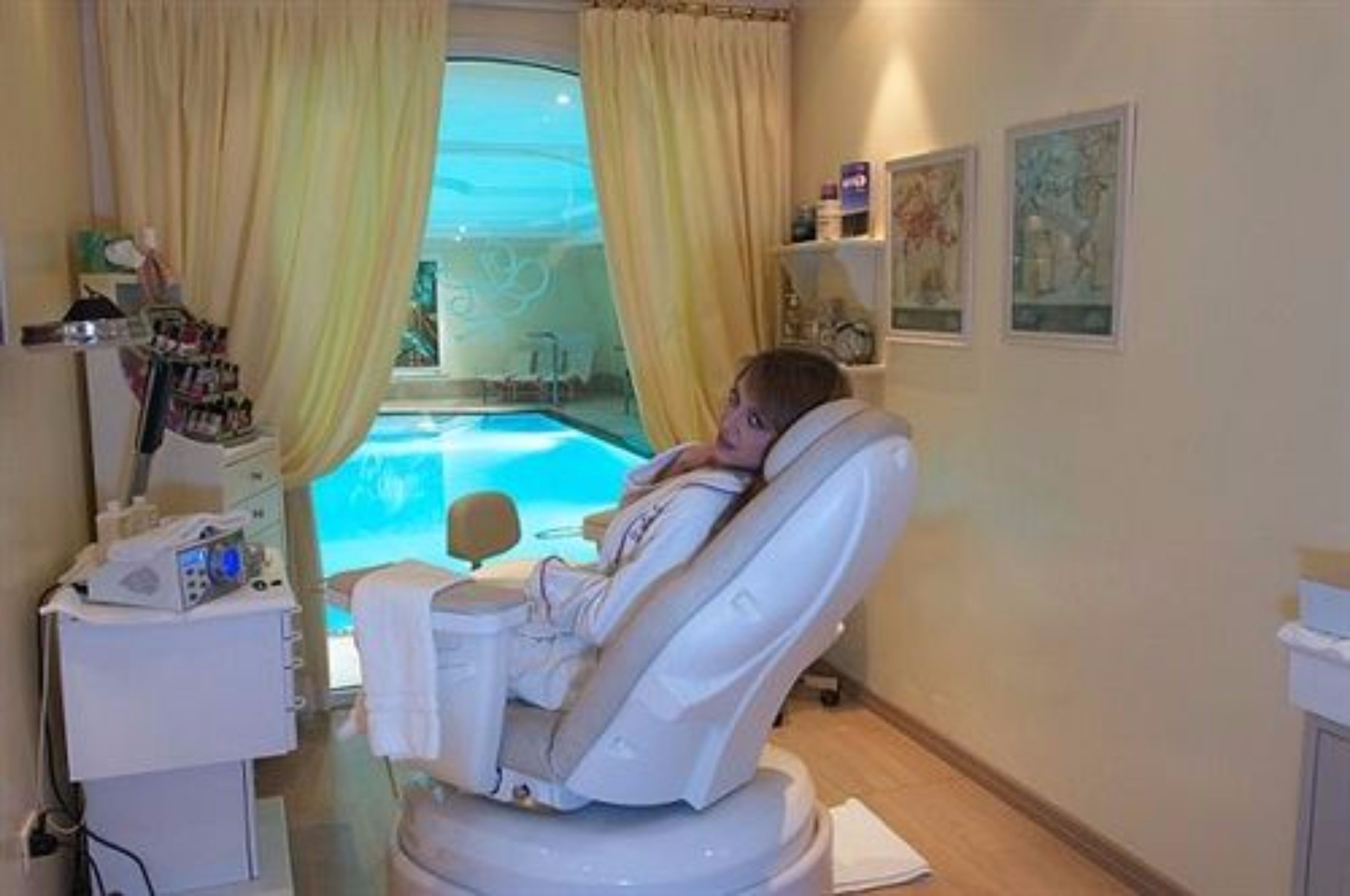 A spa room with a reclining chair, a small table with a bottle, and a window with curtains.