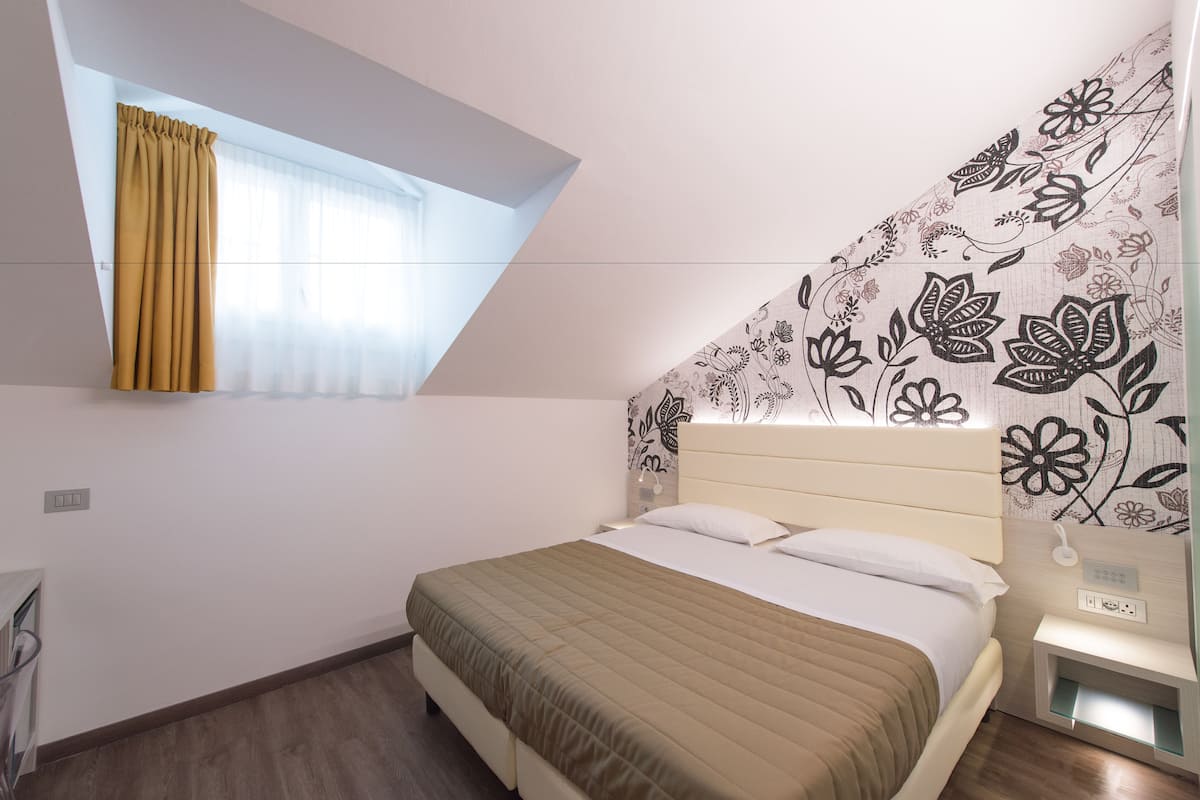 A bedroom with a bed, a window with curtains, a wall with floral wallpaper, and a bedside table with a lamp.