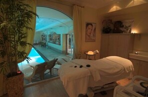 A spa room with a massage table, a small table with a lamp, and a view of an outdoor pool area. - Grand Hotel Liberty (Riva del Garda)