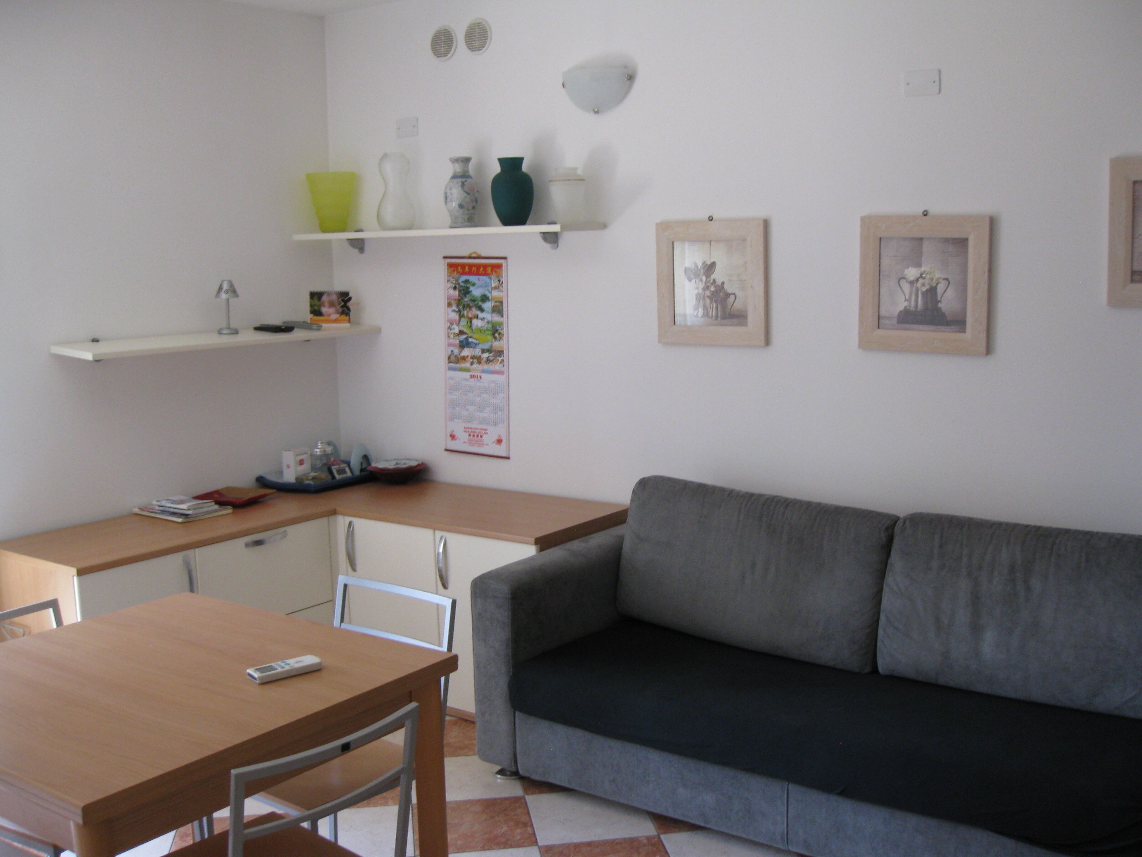 A small, well-lit room with a wooden table, a grey sofa, and a shelf with decorative items.
