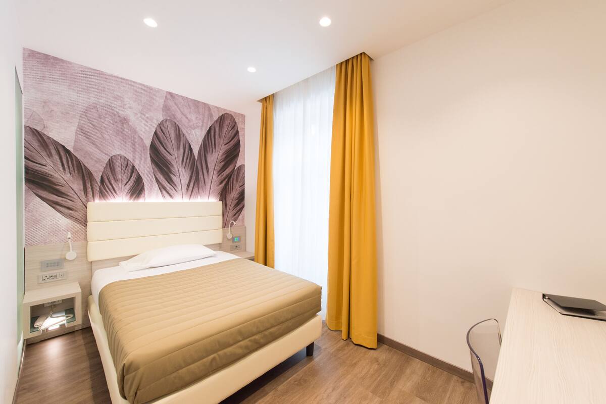 A hotel room with a bed, a nightstand, a lamp, a desk, and a large window with curtains.