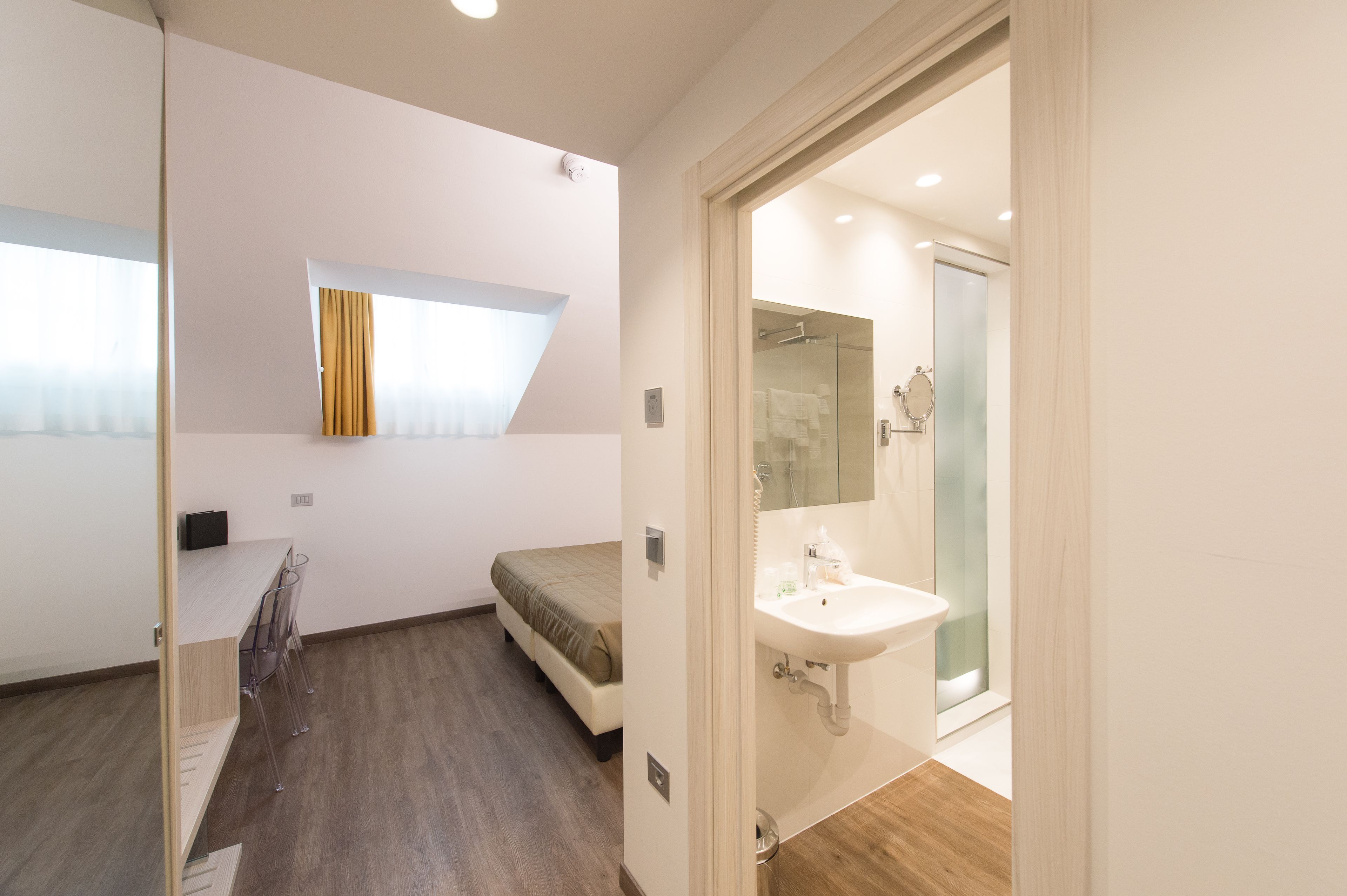 Un dormitorio moderno con una cama, una mesita pequeña y una silla. Hay un baño con lavamanos y ducha.