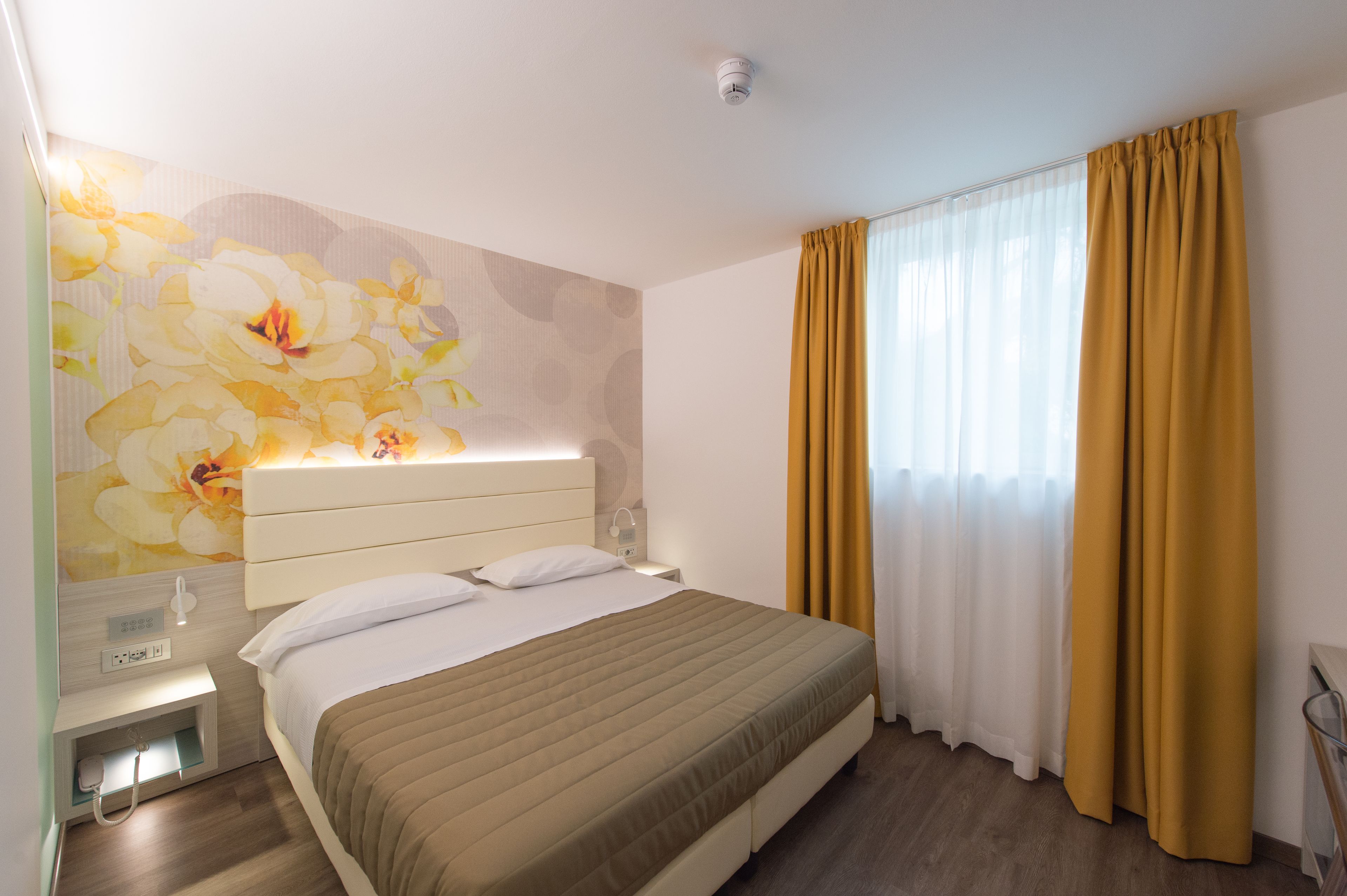 Habitación de hotel con cama, mesita de noche, lámpara de pared, enchufe y ventana con cortinas.